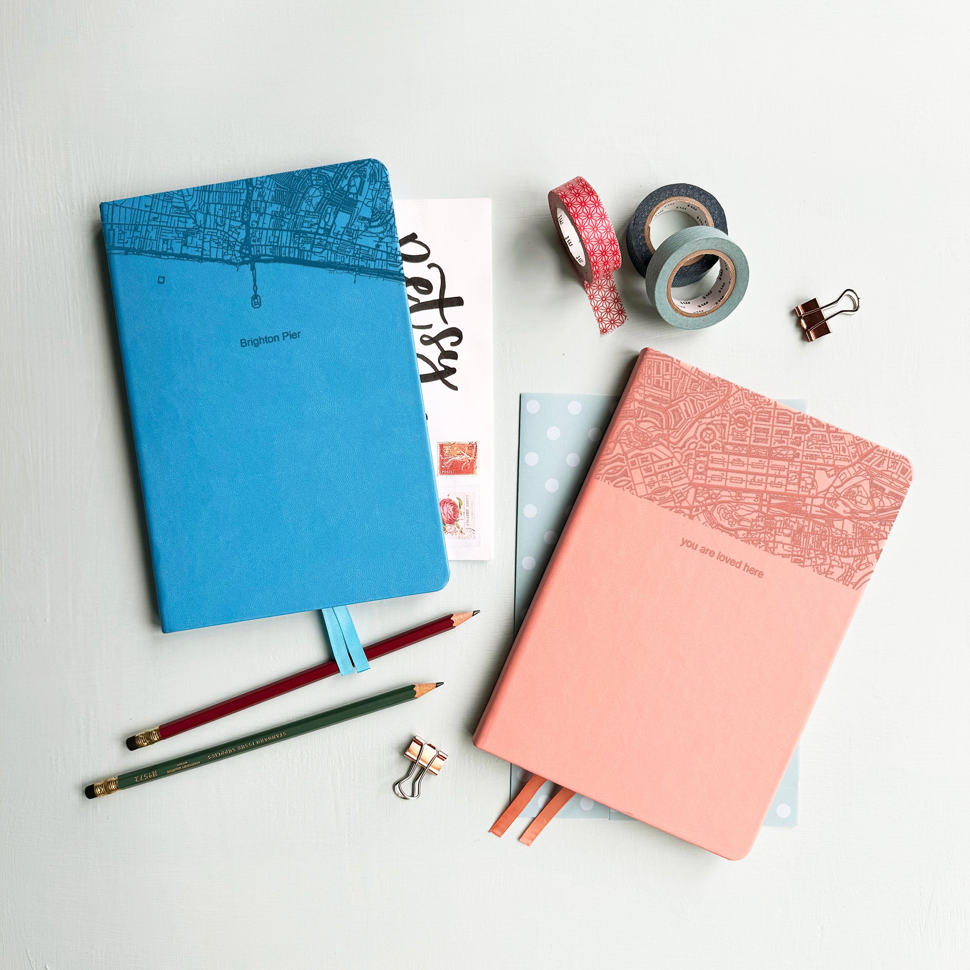 A top down image of two vegan leather notebooks, one blue and one pink. They are resting on a pale aqua table. The top third of the notebooks is engraved with a map. The font of the blue notebook is engraved with the personalised text “Brighton Pier” and The font of the pink notebook is engraved with the personalised text “you are loved here”. Surrounding the book are various stationary items.