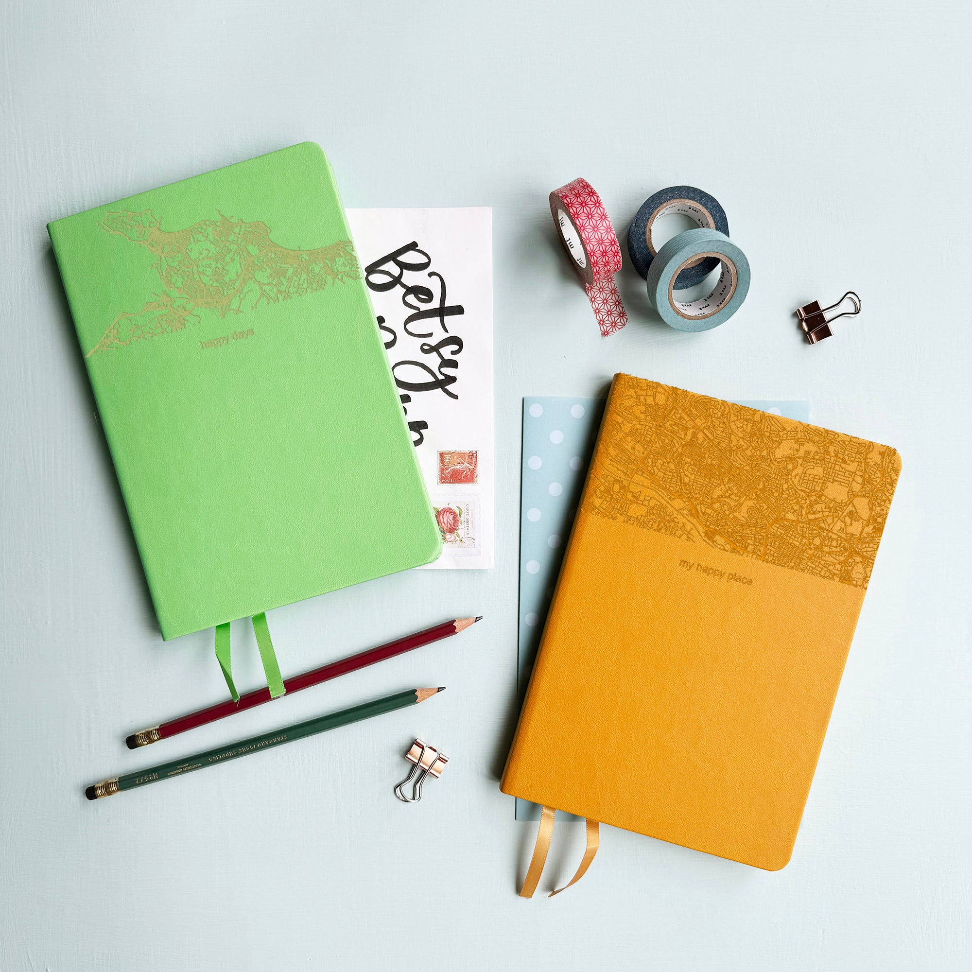 A top down image of two vegan leather notebooks, one green and one mustard yellow. They are resting on a pale aqua table. The top third of the notebooks is engraved with a map. The font of the green notebook is engraved with the personalised text “happy days” and The font of the mustard yellow notebook is engraved with the personalised text “my happy place”. Surrounding the book are various stationary items.