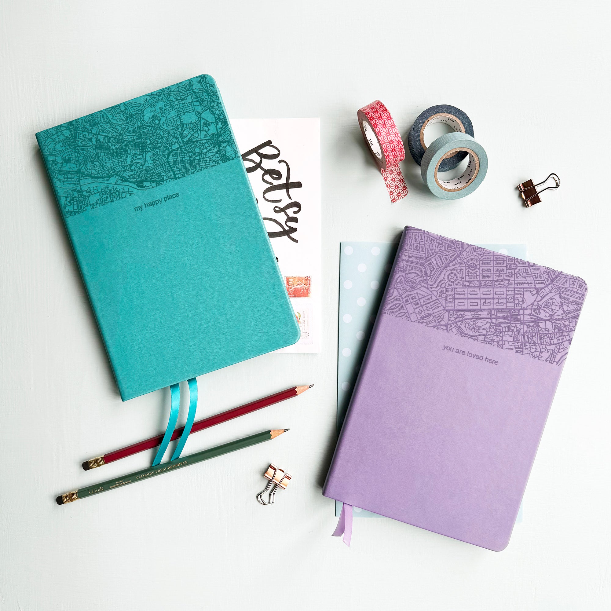 A top down image of two vegan leather notebooks, one turquoise and one lilac. They are resting on a pale aqua table. The top third of the notebooks is engraved with a map. The font of the turquoise notebook is engraved with the personalised text “my happy place” and The font of the lilac notebook is engraved with the personalised text “you are loved here”. Surrounding the book are various stationary items.