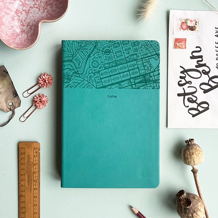 A top down image of a turquoise vegan leather notebook resting on a pale aqua table. The top third of the notebook is engraved with a map and personalised with the text “home”. Surrounding the book are various stationary items and dried flowers.