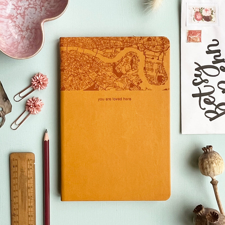 A top down image of a mustard yellow vegan leather notebook resting on a pale aqua table. The top third of the notebook is engraved with a map and personalised with the text “you are loved here”. Surrounding the book are various stationary items and dried flowers.
