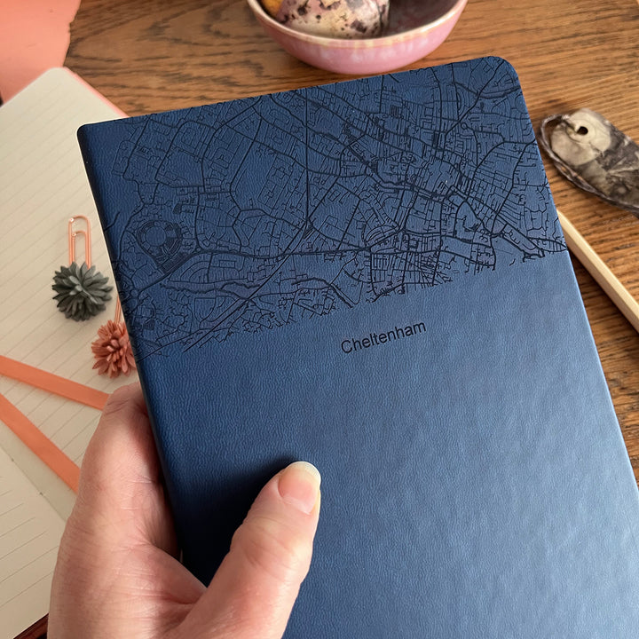A close up image of a woman's hand holding a navy blue vegan leather notebook The top third of the notebook is engraved with a map and personalised with the text “Cheltenham”. Surrounding the book are various stationary items and dried flowers resting on a brown wooden table.