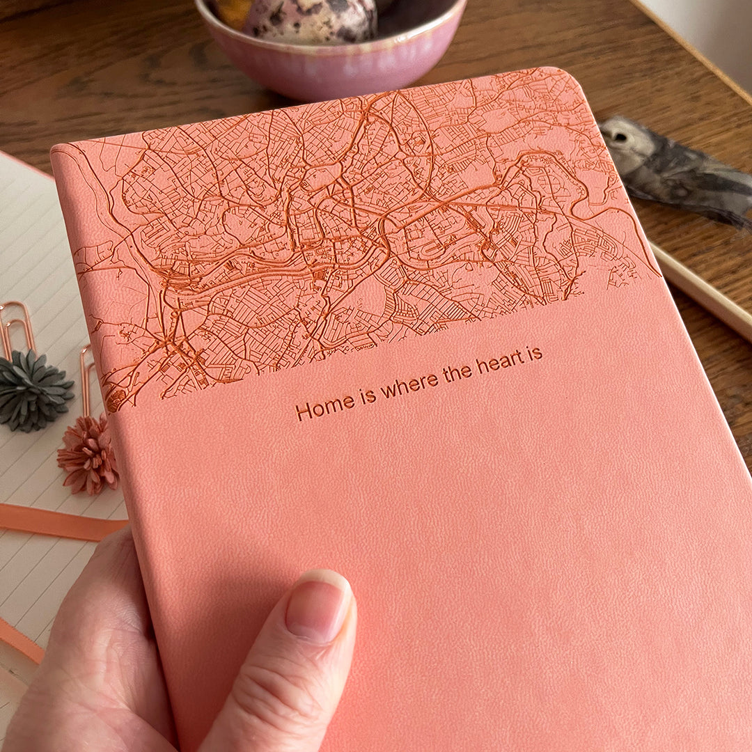 A close up image of a woman's hand holding a pink vegan leather notebook The top third of the notebook is engraved with a map and personalised with the text “home is where the heart is”. Surrounding the book are various stationary items and dried flowers resting on a brown wooden table.