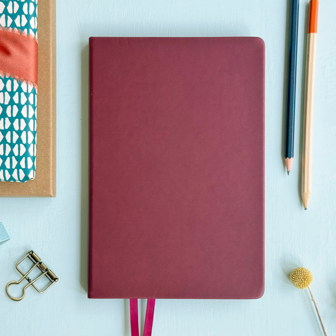 A top down image of a plain simple burgundy pink vegan leather notebook. It is resting on a light blue surface. Surrounding the book are various stationary items.