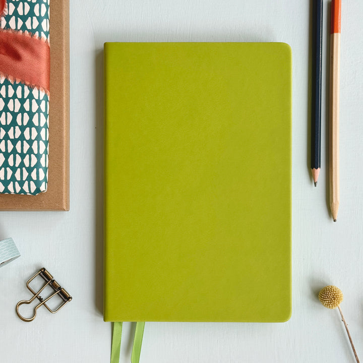 A top down image of a plain simple lime green vegan leather notebook. It is resting on a light blue surface. Surrounding the book are various stationary items.