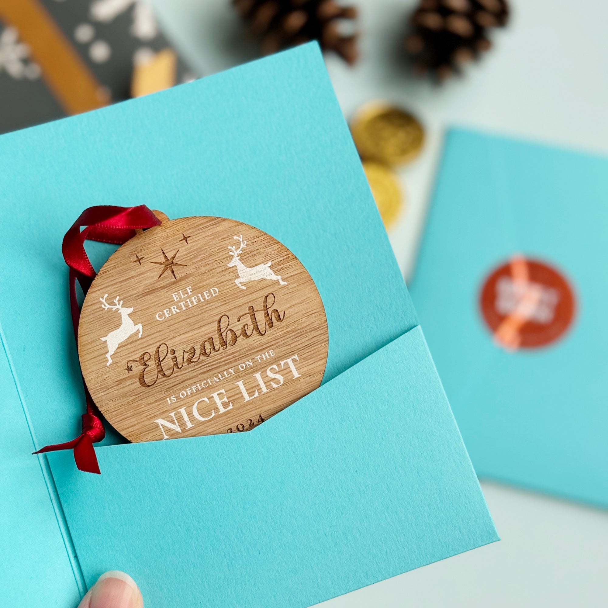 A round wooden Christmas ornament with the name "Elizabeth" engraved on it, along with the message "Elf Certified - Is Officially on the Nice List 2024" and two leaping reindeer, packaged in a blue gift box.
