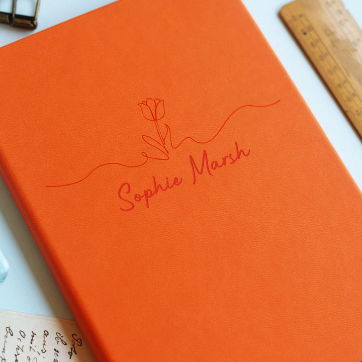 Floral motif tasteful end of year teacher gift orange notebook on pale blue background surrounded by stationery.