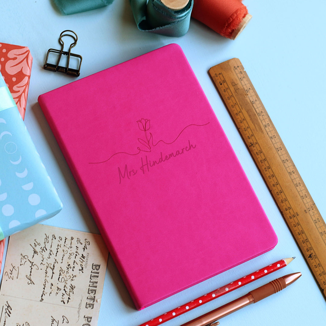 Floral motif tasteful end of year teacher gift hot pink cerise notebook on pale blue background surrounded by stationery.