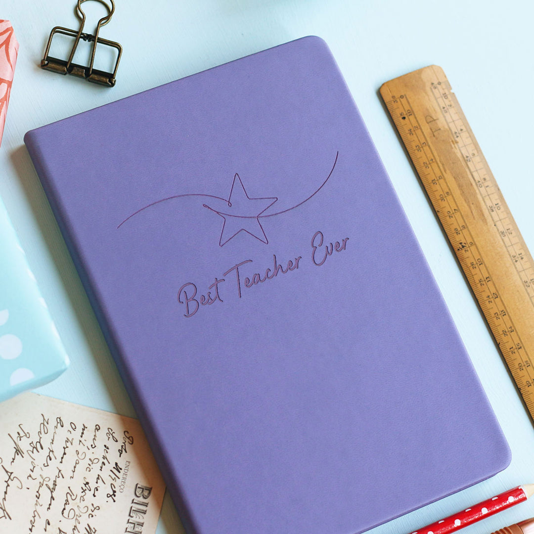 Colourful star motif fun whimsical notebook teacher gift on pale blue background, primary school best teacher ever, surrounded by stationery.