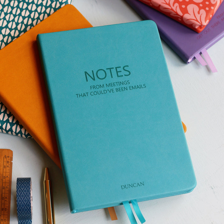 A top down image of a turquoise vegan leather notebook stacked on top of a orange notebook and resting on a pale aqua table. The front of the turquoise notebook is engraved with the phrase "Notes from meetings that could’ve been emails" and personalised with the name Duncan. Surrounding the book are various.