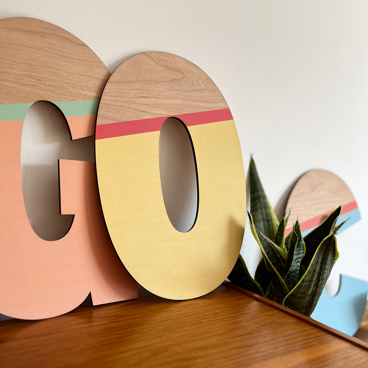 A vibrant and playful display featuring large wooden letters "G" and "O" painted in a striking color palette of peach, yellow, and green. 