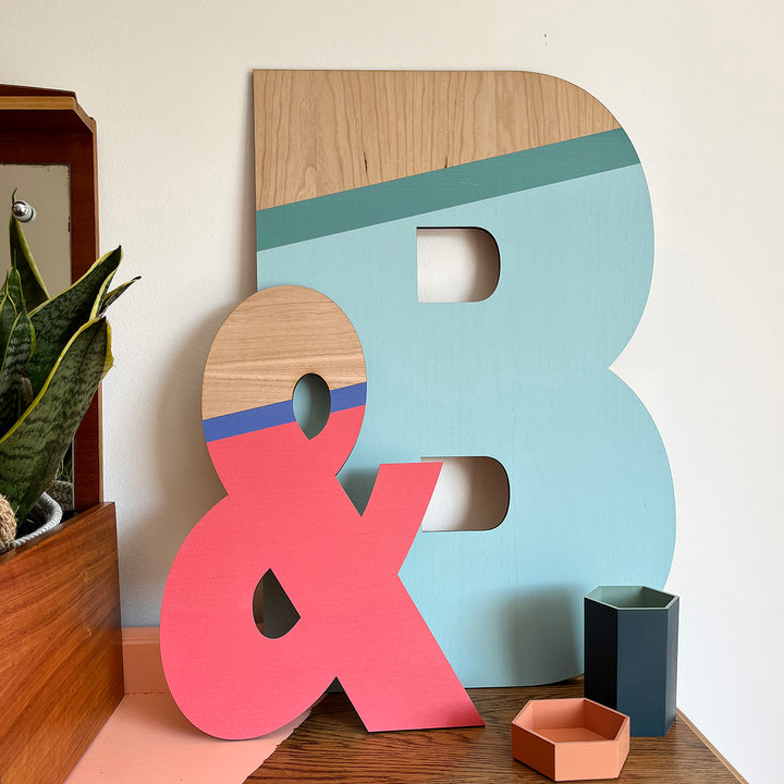 A colorful and stylish display featuring large wooden letters "B" and "&" painted in a vibrant color palette of blue, teal, and pink. The letters are showcased on a shelf alongside a plant and other decorative items, adding a playful touch to the space.