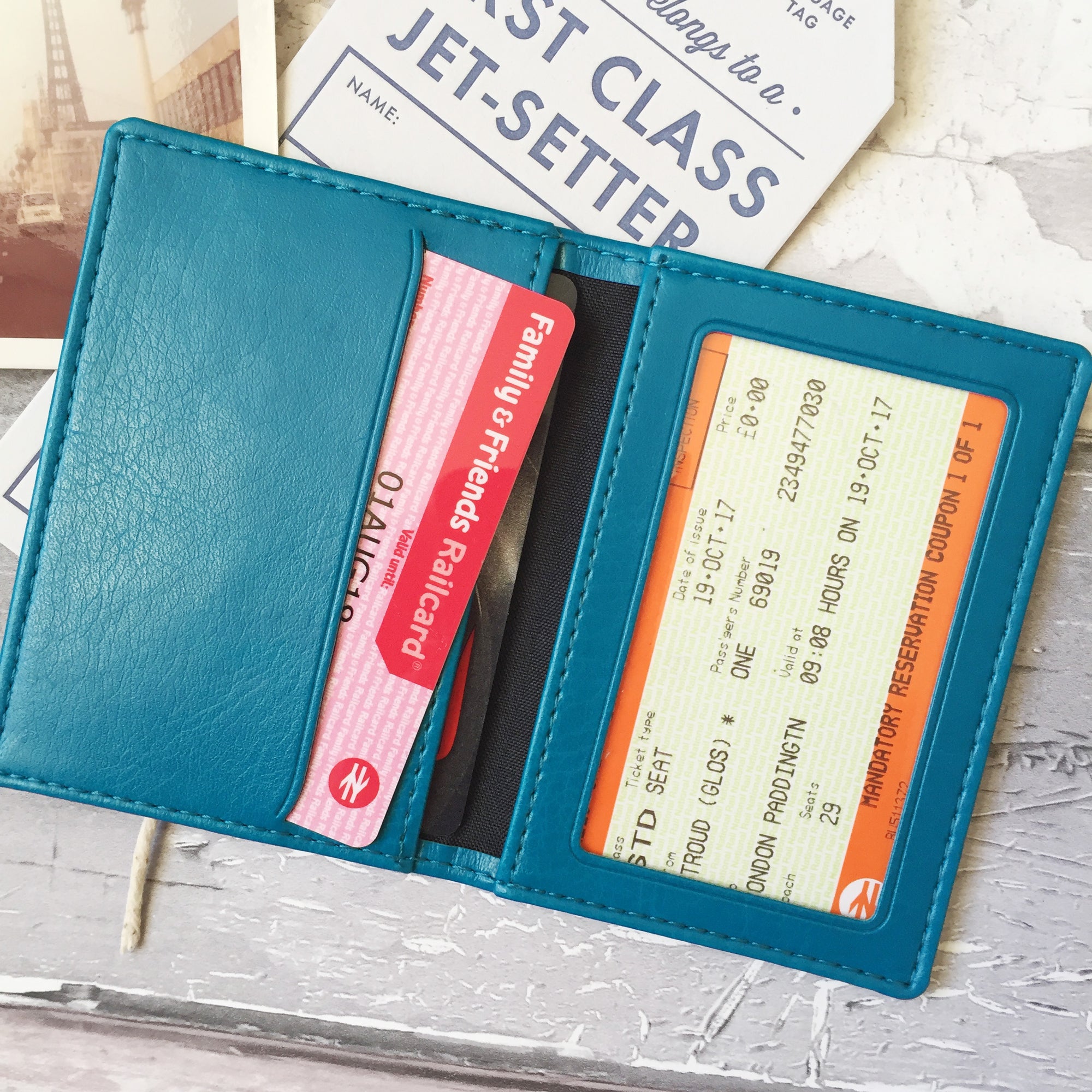 A womans hand holds a turquoise custom ID holder/bus pass leather holder. The front of the slim style wallet has been personalised with the words Emergency contacts and then three mobile phone numbers.