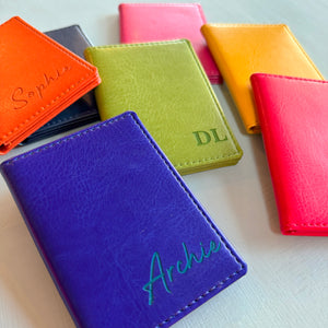 A collection of the colours available of the slim wallet bus pass holder. Showing the leather car holder in airforce blue, royal blue, red, orange, yellow, pink, and green. Some have been engraved with names or initials in the bottom right hand corner