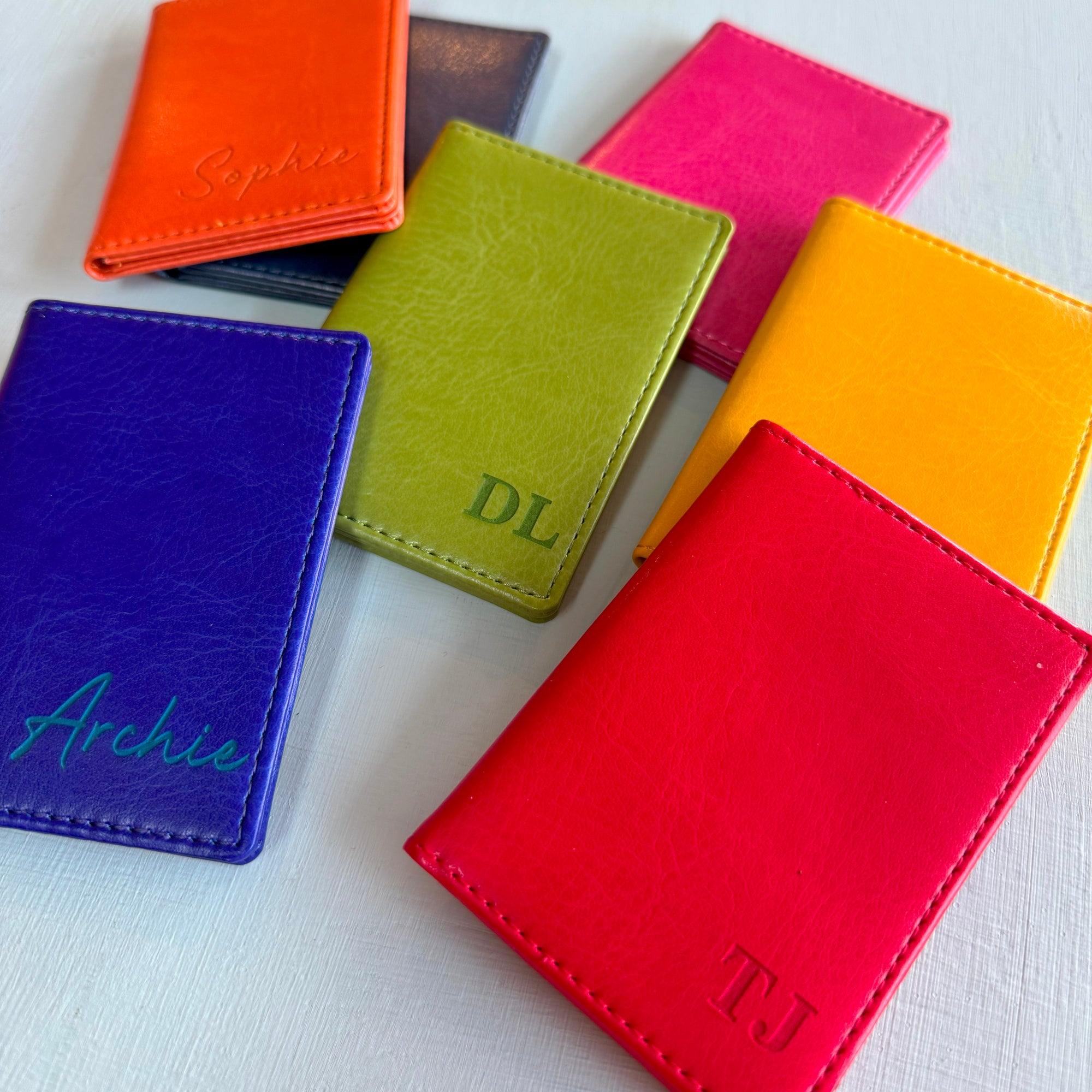 A collection of the colours available of the slim wallet bus pass holder. Showing the leather car holder in airforce blue, royal blue, red, orange, yellow, pink, and green. Some have been engraved with names or initials in the bottom right hand corner