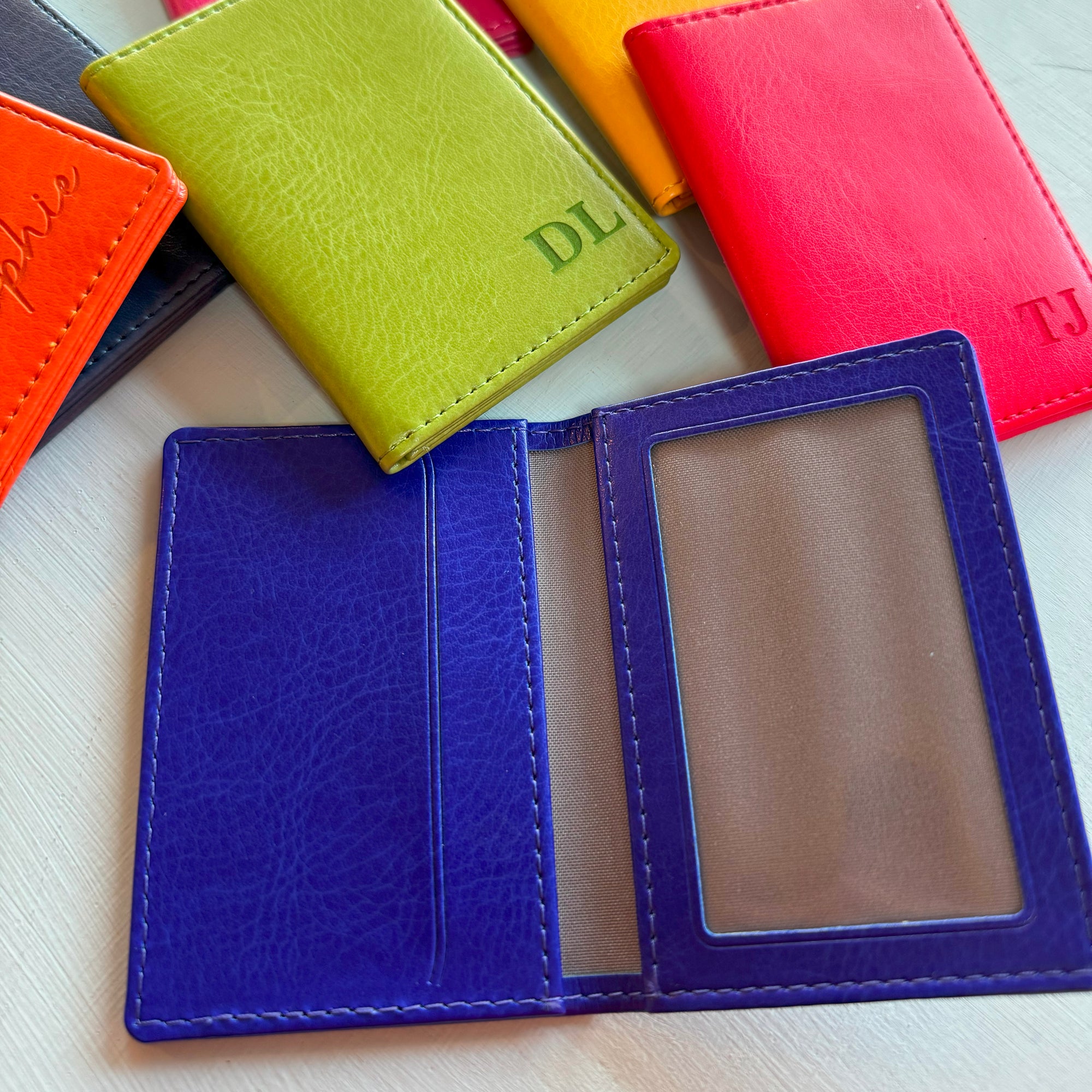 A collection of the colours available of the slim wallet bus pass holder. Showing the leather card holder in airforce blue, royal blue, red, orange, yellow, pink, and green. Some have been engraved with names or initials in the bottom right hand corner. The royal blue car holder is open, clearly showing the transparent side for a photocard and the opposite side for credit cards.
