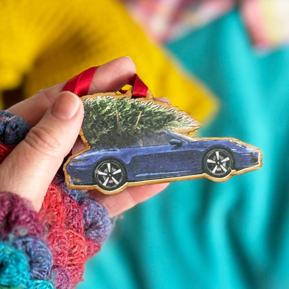 A hand holding a wooden car enthusiasts gift Porsche 911 Christmas tree decoration with a blue car and green tree on a blue and yellow background.