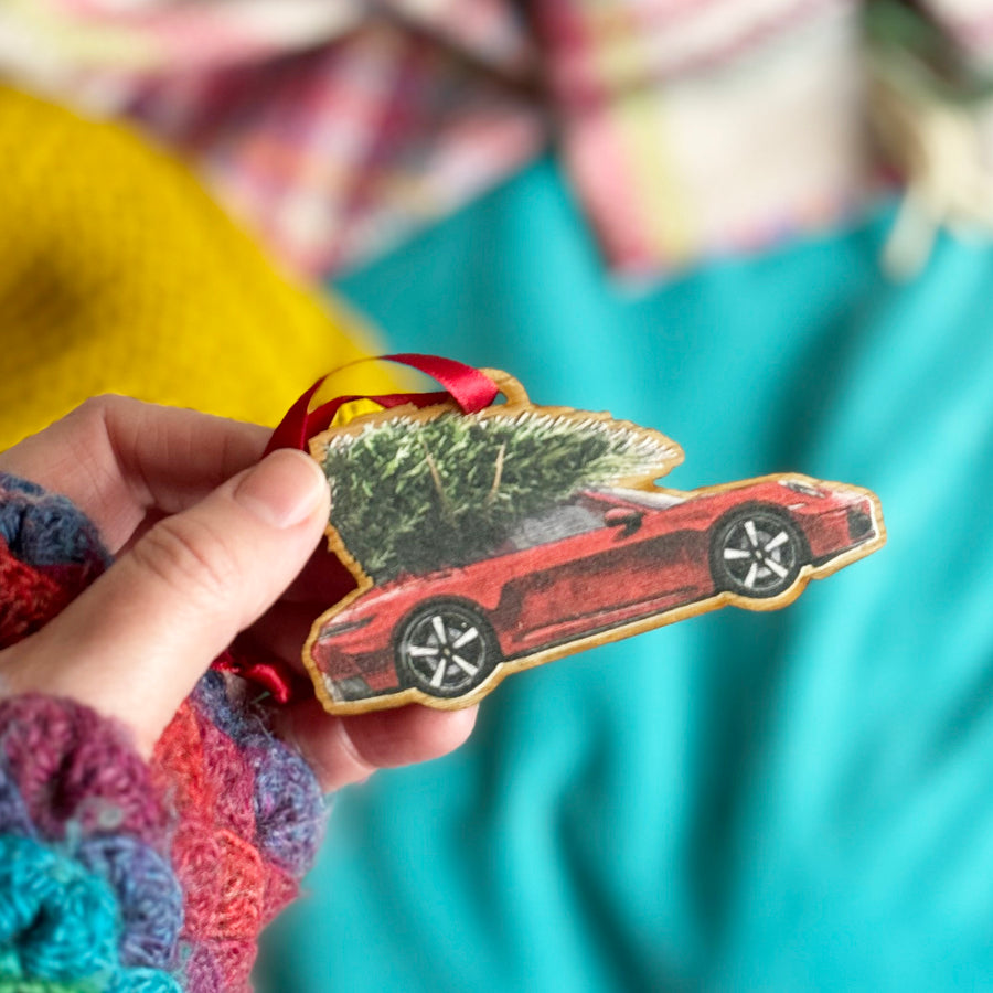 A hand holding a wooden car enthusiasts gift Porsche 911 Christmas tree decoration with a red car and green tree on a blue and yellow background.