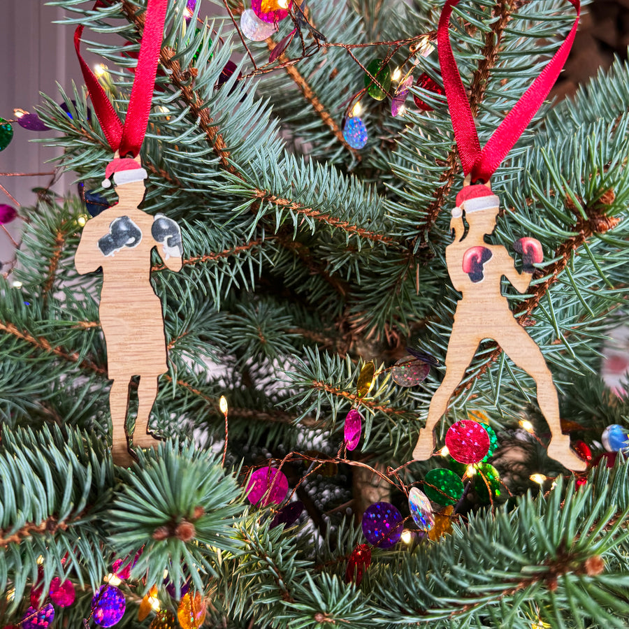 Two wooden Christmas tree gift ornaments depicting silhouettes of a boxer and a yoga practitioner wearing Santa hats, hanging from the branches of a decorated Christmas tree.