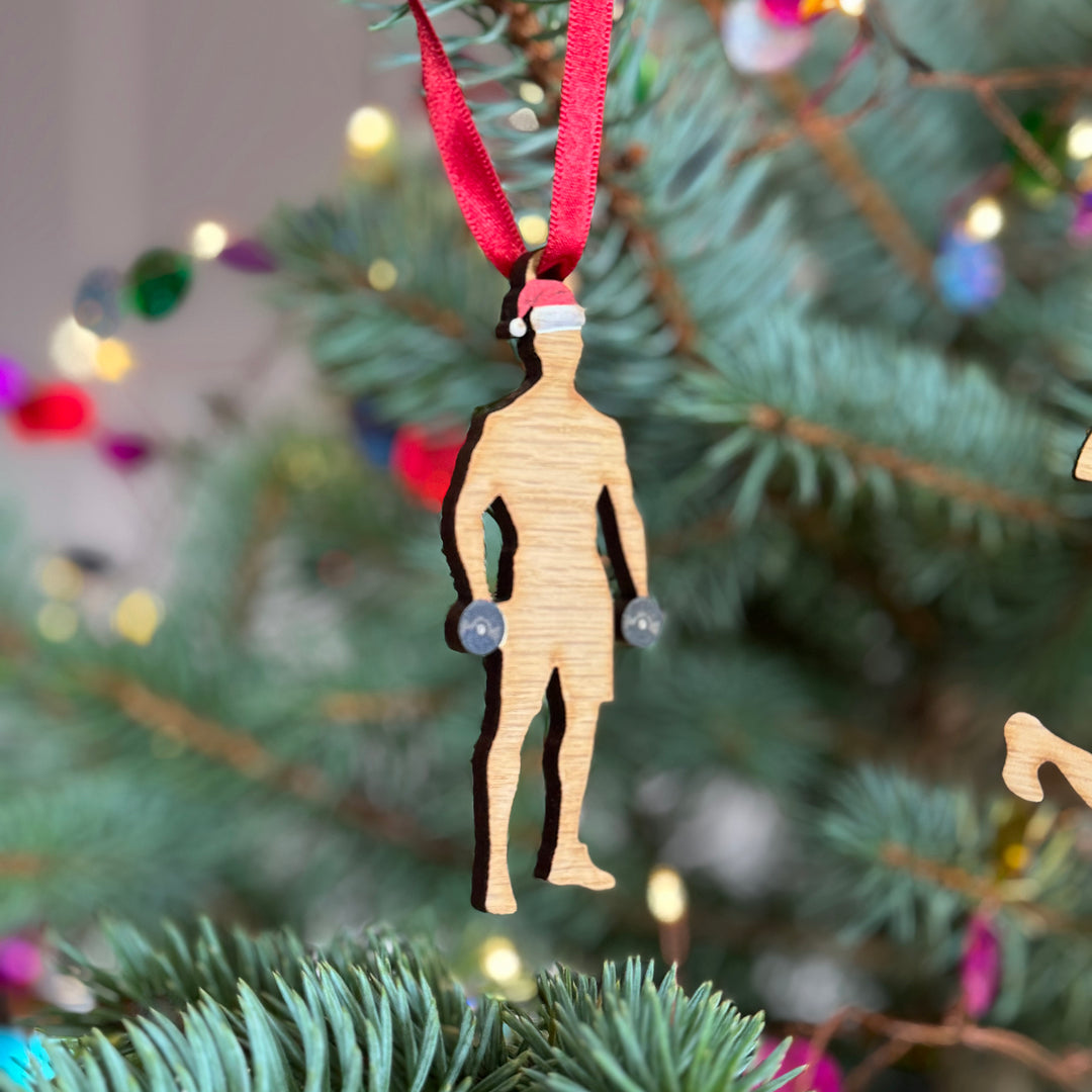 Weightlifter Christmas tree decoration ornament for sports lover, wooden silhouette made from sustainable cherry veneer, printed with a festive hat and a set of weights, hung from a red ribbon on a tree.