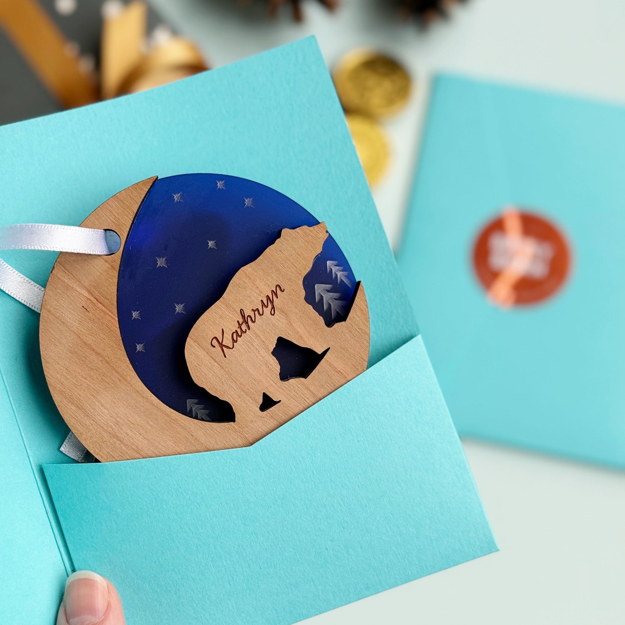 A hand holds a teal card with a round wooden ornament featuring a bear cutout and the name "Kathryn." The card is open, revealing a dark blue inset with stars and other cards and pine cones in the background.