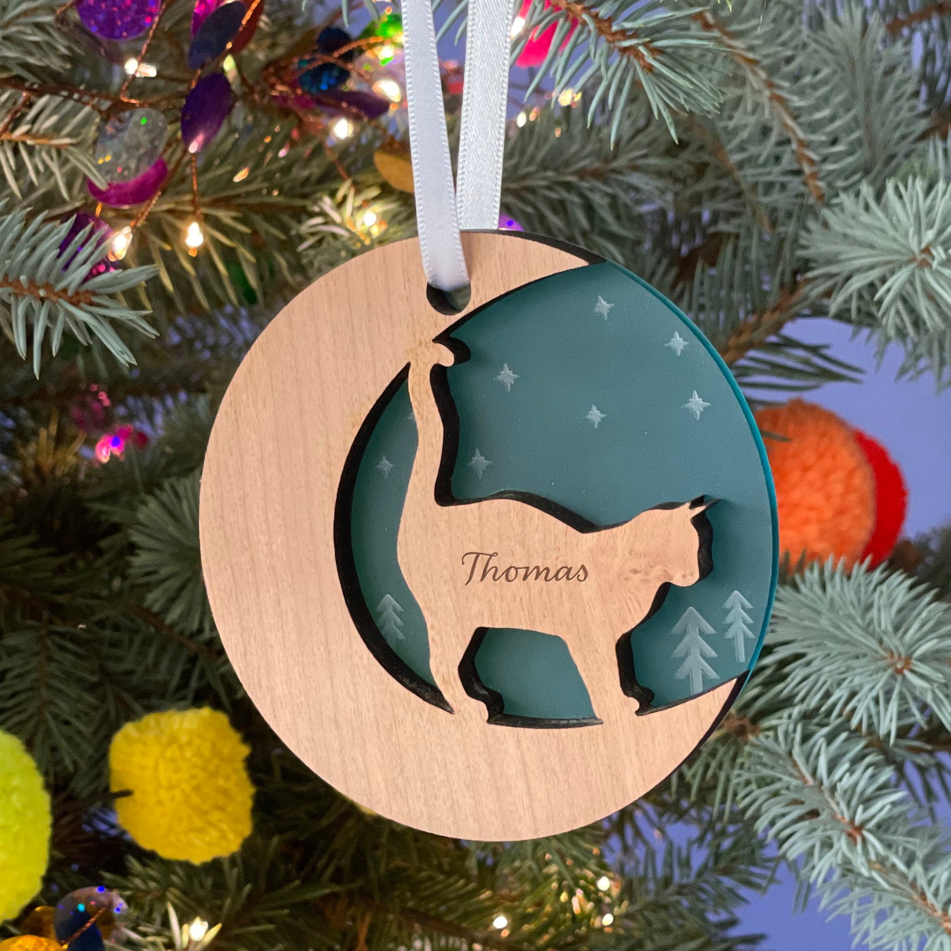A round wooden Christmas ornament hangs on a pine branch against a blurred background with colorful decorations. The ornament features a cutout silhouette of a cat and is engraved with the name "Thomas."