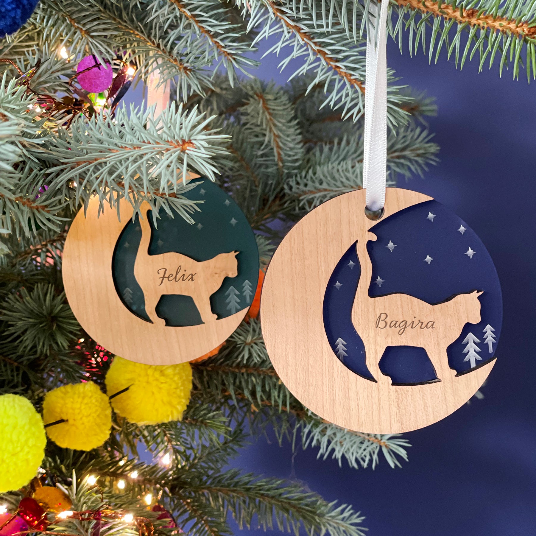 Two round wooden Christmas ornaments hang on a pine branch against a dark blue background. Each ornament features a cutout silhouette of a cat and is engraved with a name, "Felix" and "Bagira." Colorful decorations are visible on the branch.