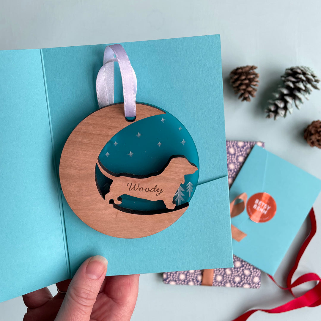 A hand holds a teal card with a round wooden ornament featuring a dachshund cutout and the name "Woody." The card is open, revealing a patterned interior and other cards with pine cones in the background.