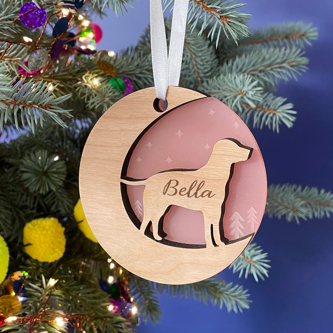 A round wooden Christmas ornament hangs on a pine branch against a dark blue background. The ornament features a cutout silhouette of a dog and is engraved with the name "Bella." The inset is a light pink color.