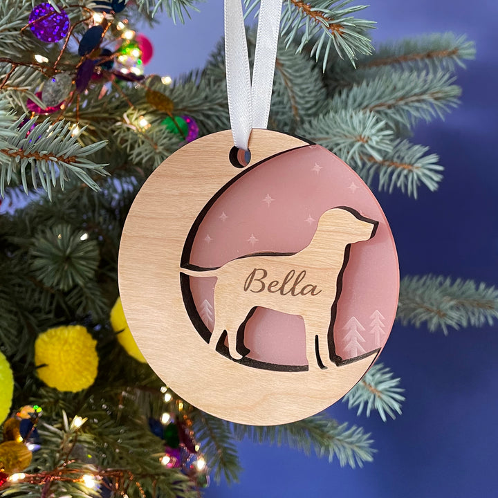 A round wooden Christmas ornament hangs on a pine branch against a dark blue background. The ornament features a cutout silhouette of a dog and is engraved with the name "Bella." The inset is a light pink color.