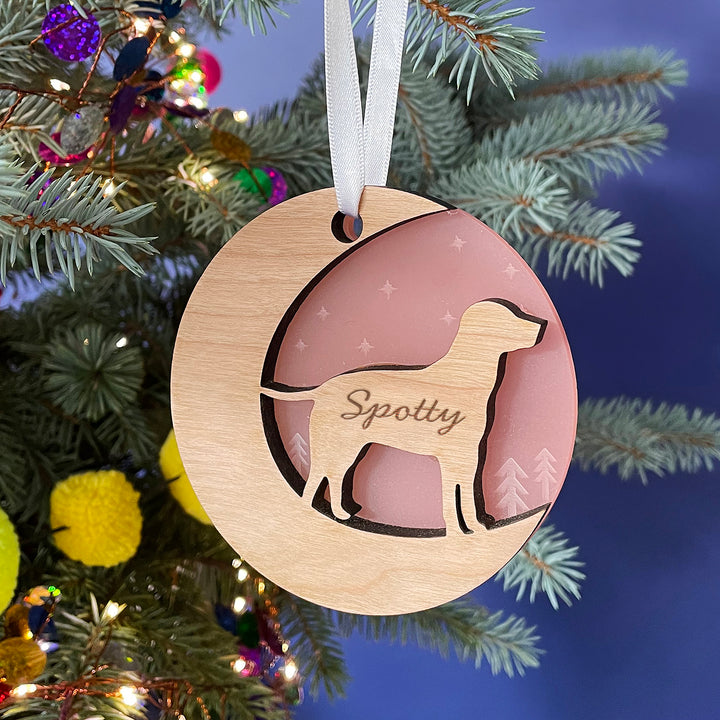 A round wooden Christmas ornament hangs on a pine branch against a dark blue background. The ornament features a cutout silhouette of a dog and is engraved with the name "Spotty." The inset is a light pink color.
