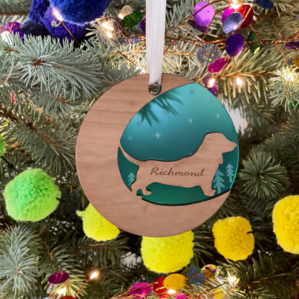 A round wooden Christmas ornament hangs on a pine branch against a blurred background with colorful pom-poms. The ornament features a cutout silhouette of a dachshund and is engraved with the name "Richmond."