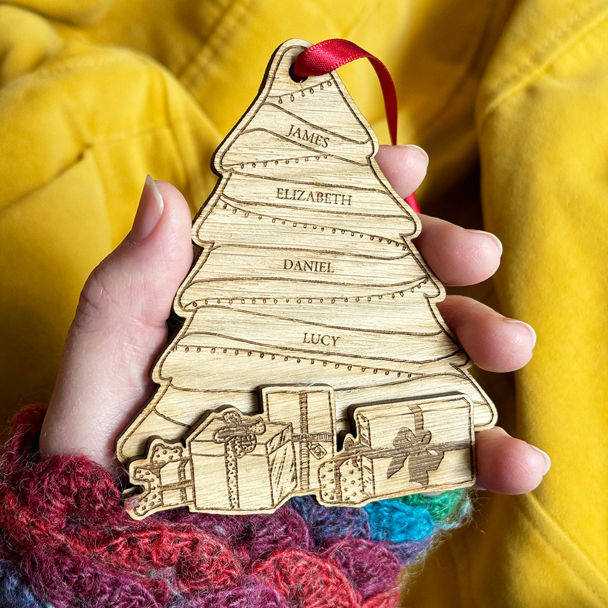 A personalized wooden Christmas tree ornament with engraved names, held in a hand with a red ribbon loop, against a yellow fabric background.