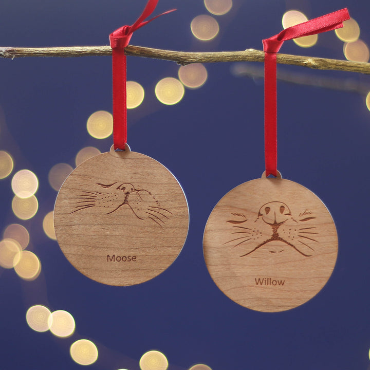 Eco pet lovers Christmas gift on two sustainable cherry veneered wood Xmas tree ornaments with engraved faces and personalised cat or dog name, strung with red ribbons against a festive background.
