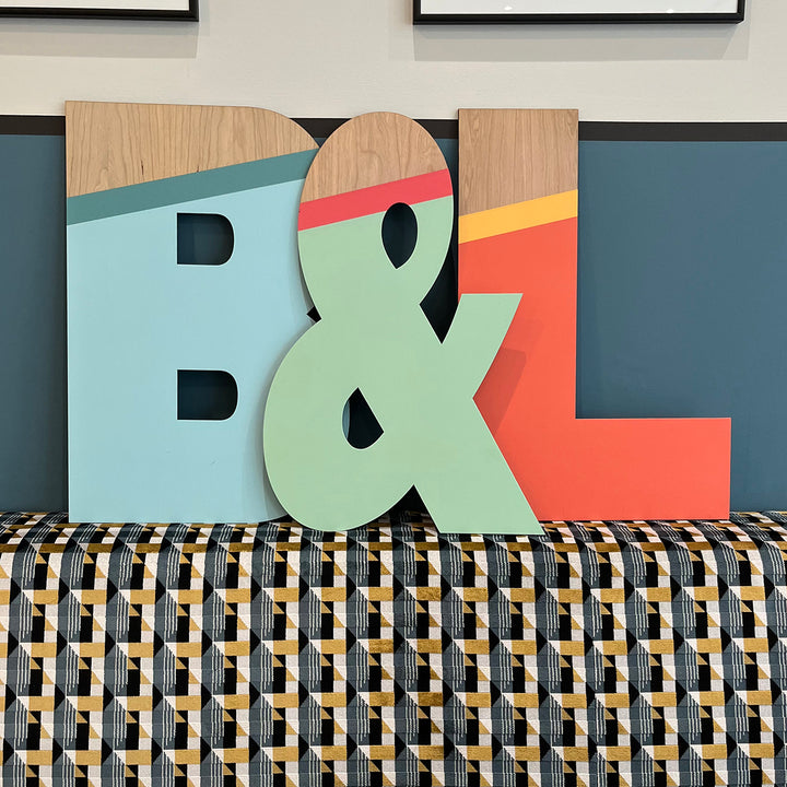 Large wooden letters "B" and "L" painted in a vibrant color scheme, with an ampersand in between, displayed on a table with a patterned tablecloth.

