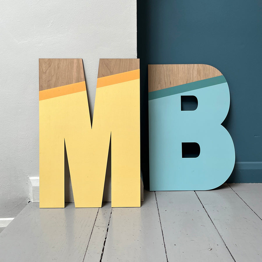 Large wooden letters "M" and "B" with pastel yellow and blue tones, featuring a natural wood accent, leaning against a two-tone wall.