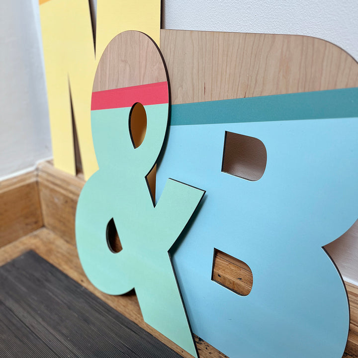 A vibrant and playful display featuring large wooden letters "M," "&," and "B" painted in a striking color palette of yellow, green, blue, and natural wood tones. 