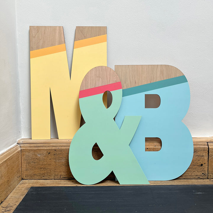 Large wooden letters "M," "&," and "B" painted in vibrant color palette of yellow, green, blue, and natural wood tones, displayed on a floor.


