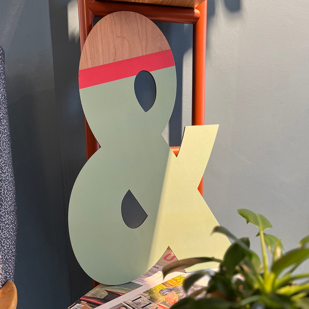 A large wooden ampersand painted in shades of green with a red stripe, leaning against a chair in a room.
