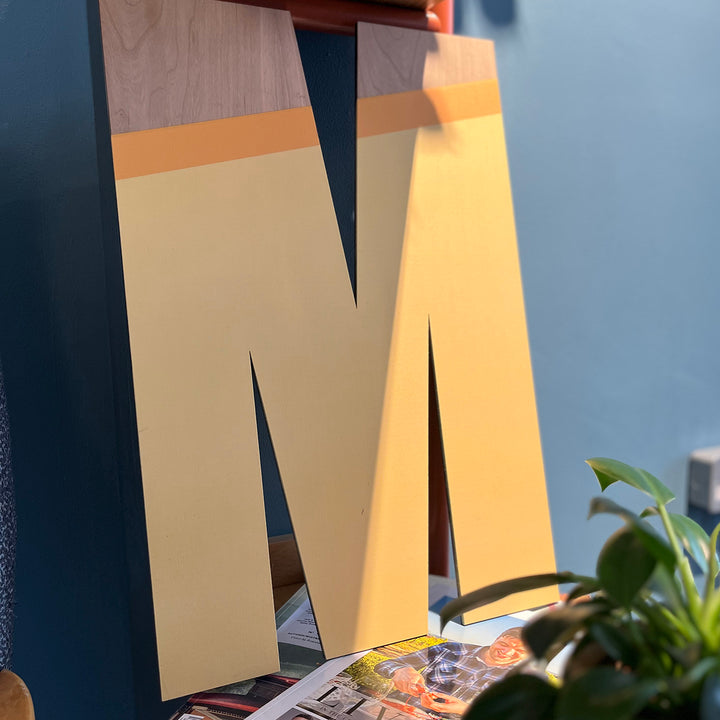 A large wooden letter "M" painted in a warm yellow with a contrasting wood grain detail, leaning against a wall.
