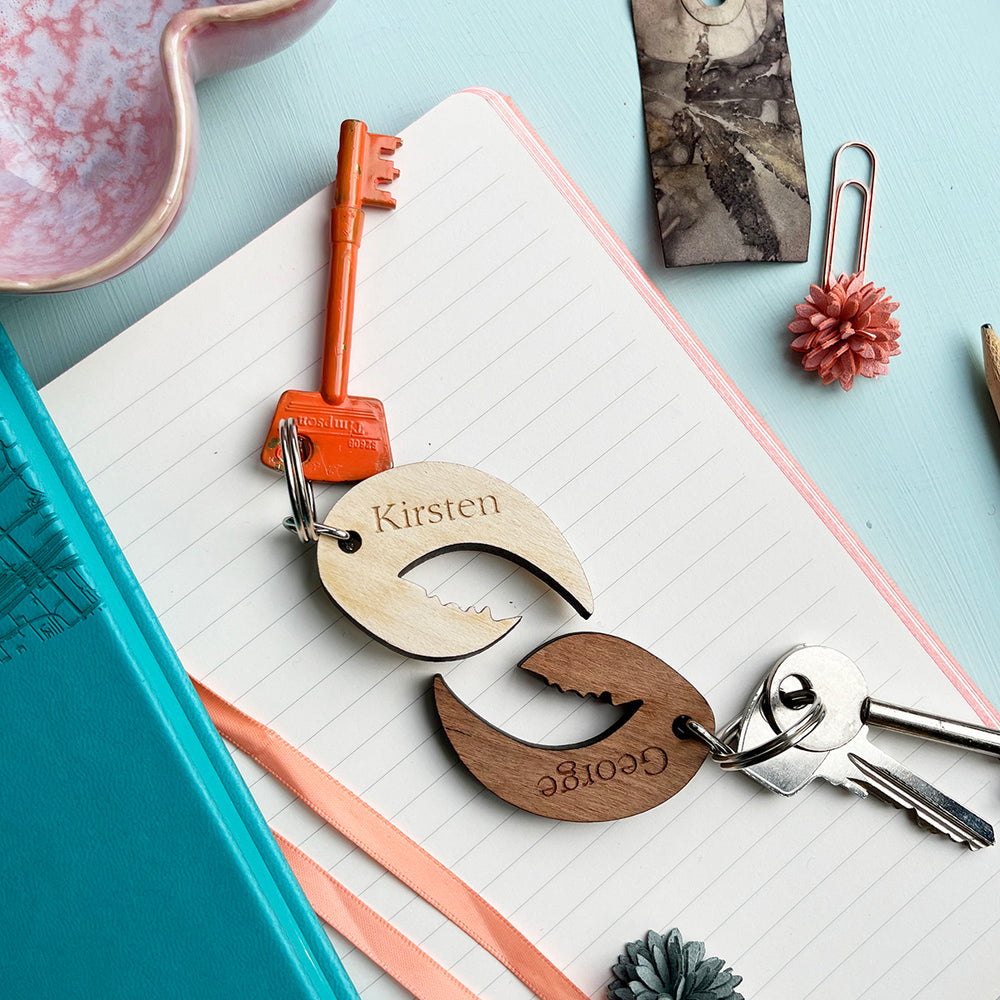Two wooden lobster claw keyrings with the names "Kirsten" and "George" engraved on them, lying on a notebook with other stationery items.