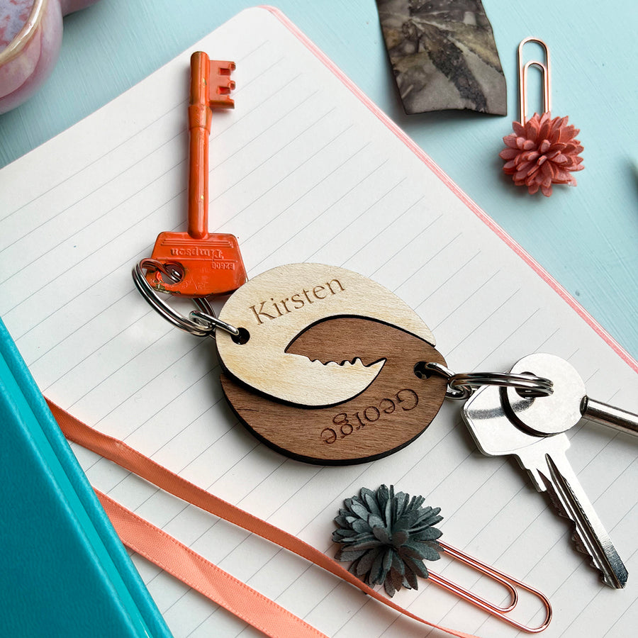 A charming pair of personalized keyrings, crafted from wood and designed to fit together like puzzle pieces. The keyrings are engraved with the names "Kirsten" and "George," creating a unique and thoughtful gift for a couple.
