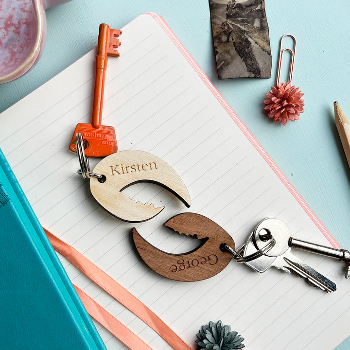 Two wooden lobster claw keyrings with the names "Kirsten" and "George" engraved on them, lying on a notebook with other stationery items.