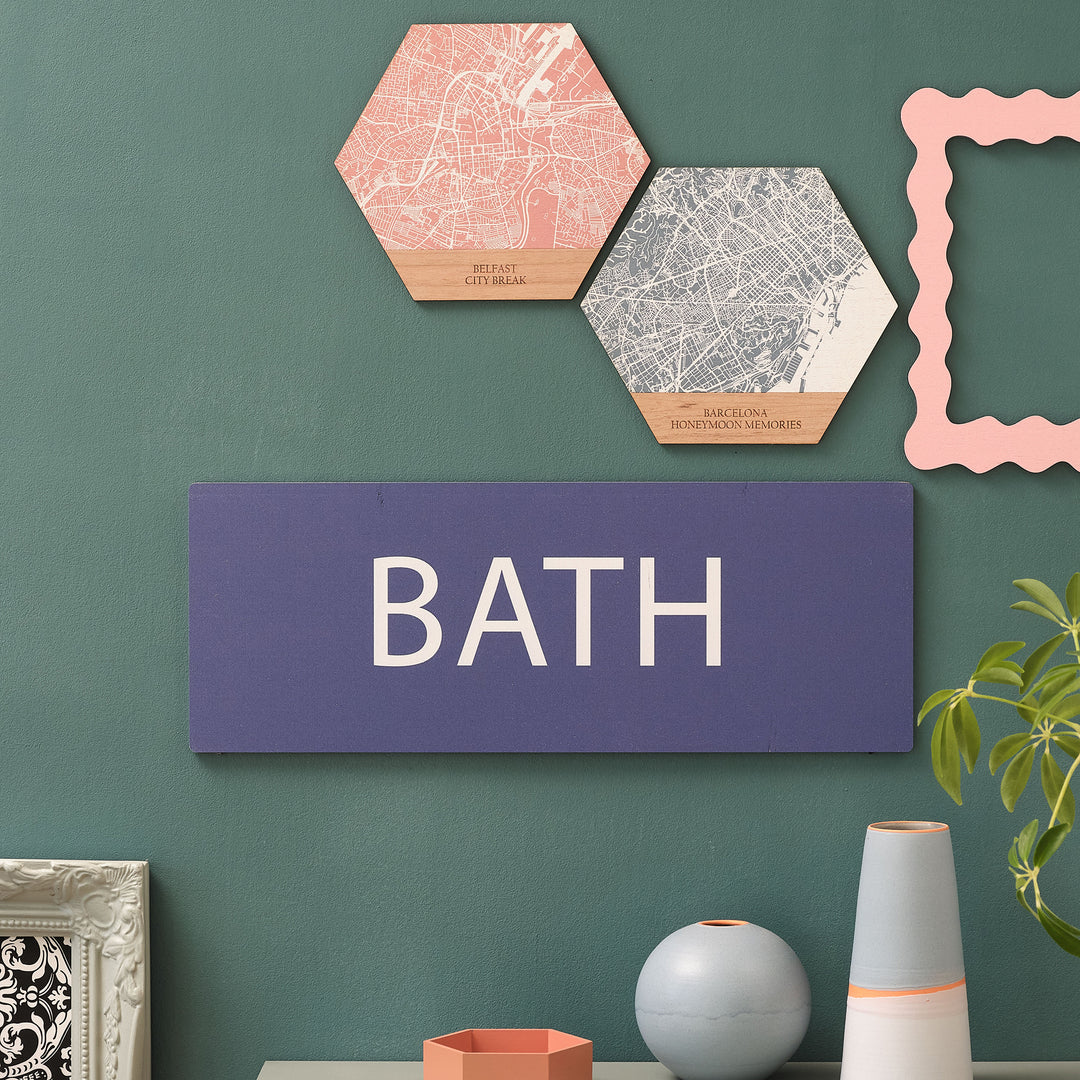 Royal blue rectangle wooden wall sign with personalised location travel memory 'BATH' text in stylish white letters. Hung on a forest green wall with picture frames and ceramics on a shelf below.