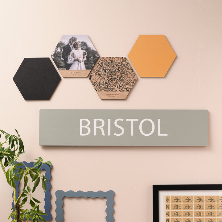 Sleek modern wooden rectangular destination wall print of 'BRISTOL' pale white text on a sage green print. On a pale peach wall with other wall prints and plants around.