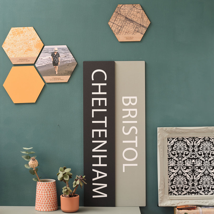 A wooden sign with the text 'Cheltenham' printed in large black letters, displayed vertically. The sign is placed on a wall with other decorative items and a map in the background. Next to it is a pale blue travel memory wooden plaque sign with 'Bristol' printed on it. Choose your own destination and colour to personalise this elegant wall print.