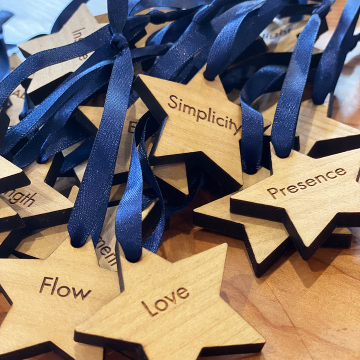 Wooden star ornaments with engraved words like "Simplicity," "Presence," "Flow," and "Love," tied with navy blue ribbons.