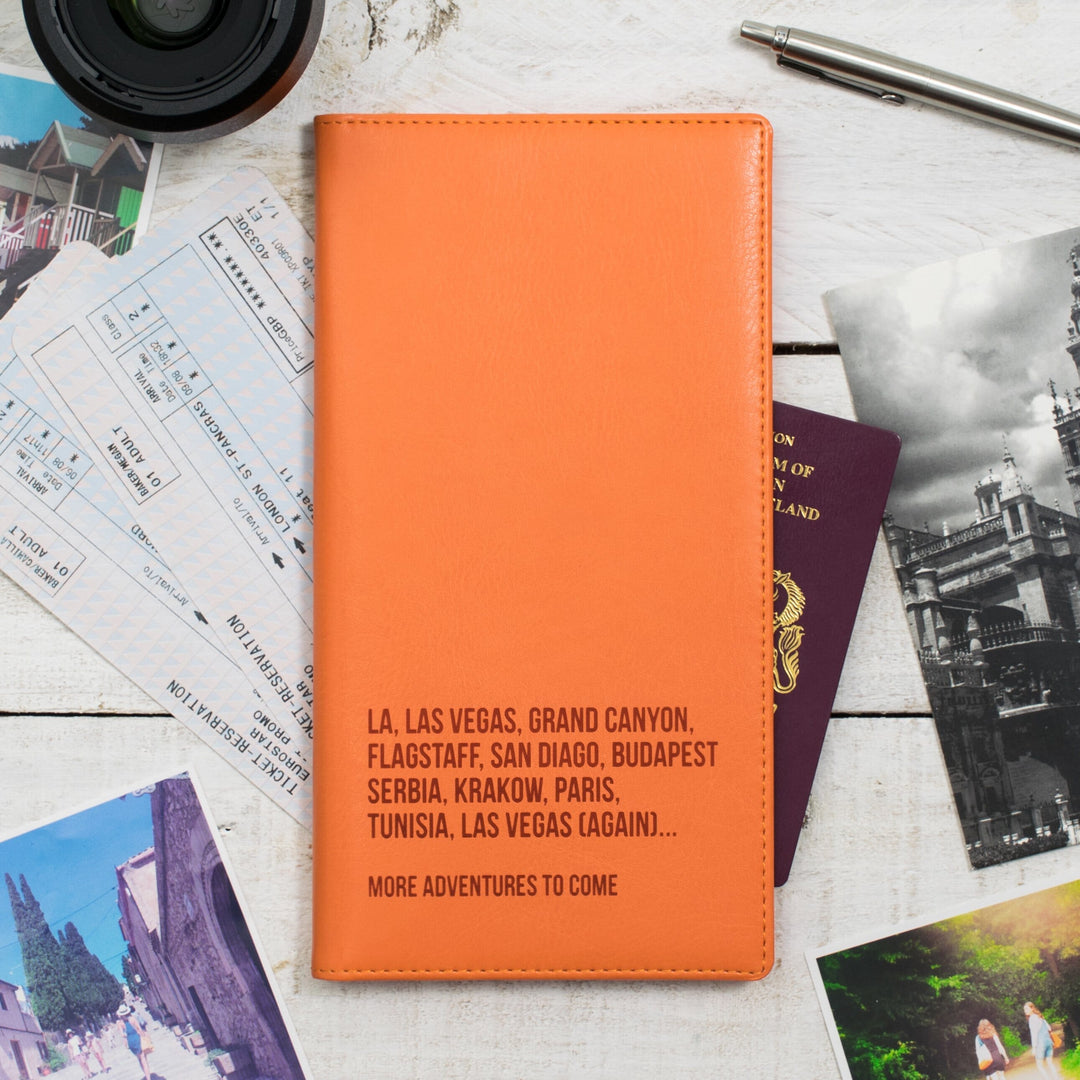 A orange personalized travel wallet with destination names embossed on it, placed on a wooden surface alongside passports, boarding passes, travel photos, and a pen.