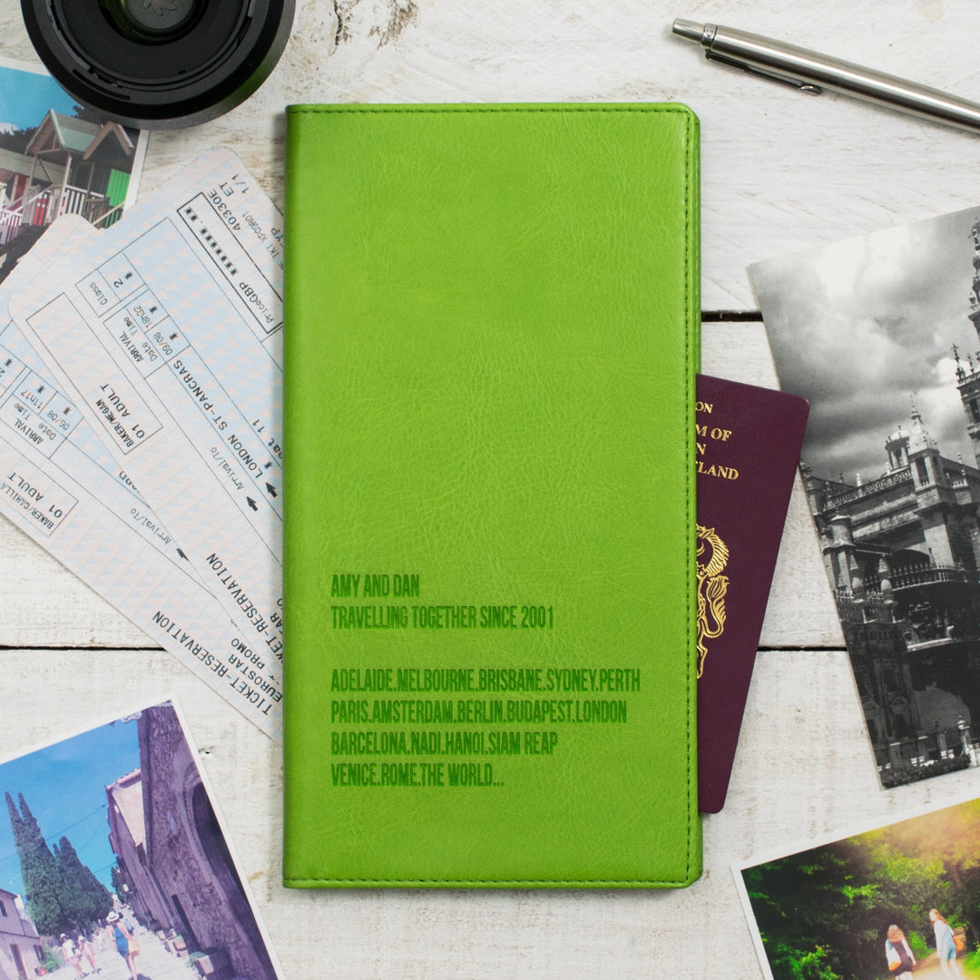 A green personalized travel wallet with destination names embossed on it, placed on a wooden surface alongside passports, boarding passes, travel photos, and a pen.