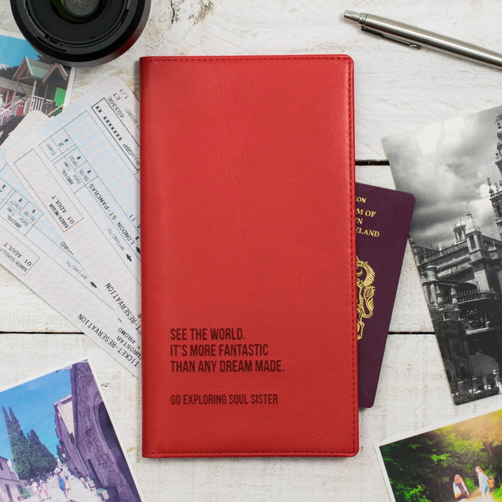 A red personalized travel wallet with destination names embossed on it, placed on a wooden surface alongside passports, boarding passes, travel photos, and a pen.